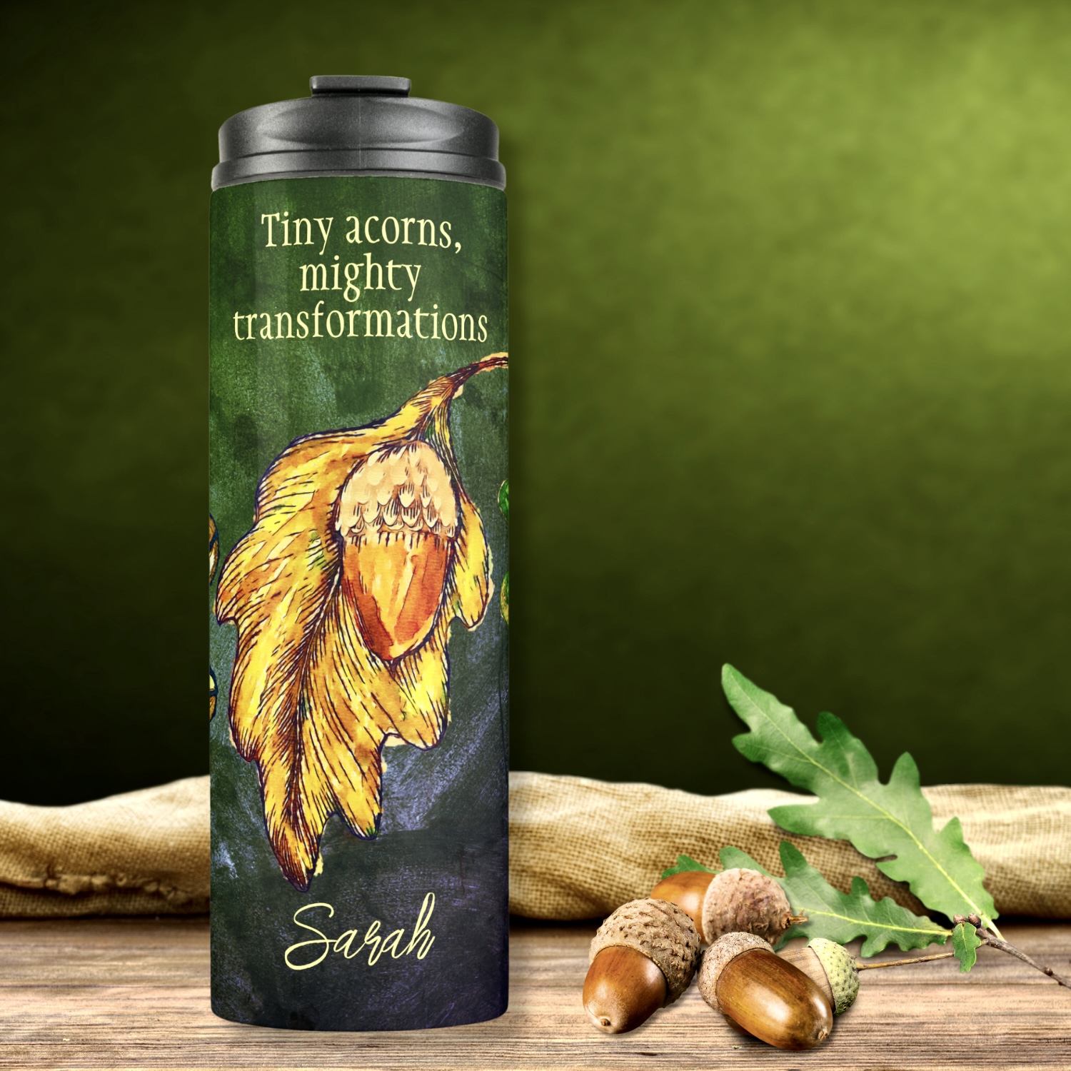 Acorn Fall Colors Inspirational Thermal Tumbler with lime and golden dry leaves, brightly painted acorns in golden and orange, arranged as a collage on a dark hunter green washed out surface. Bold design featuring the quote "Tiny acorns, mighty transformations." Customizable with your favorite quote, phrase, or personalized name. Two custom fields for personalization.