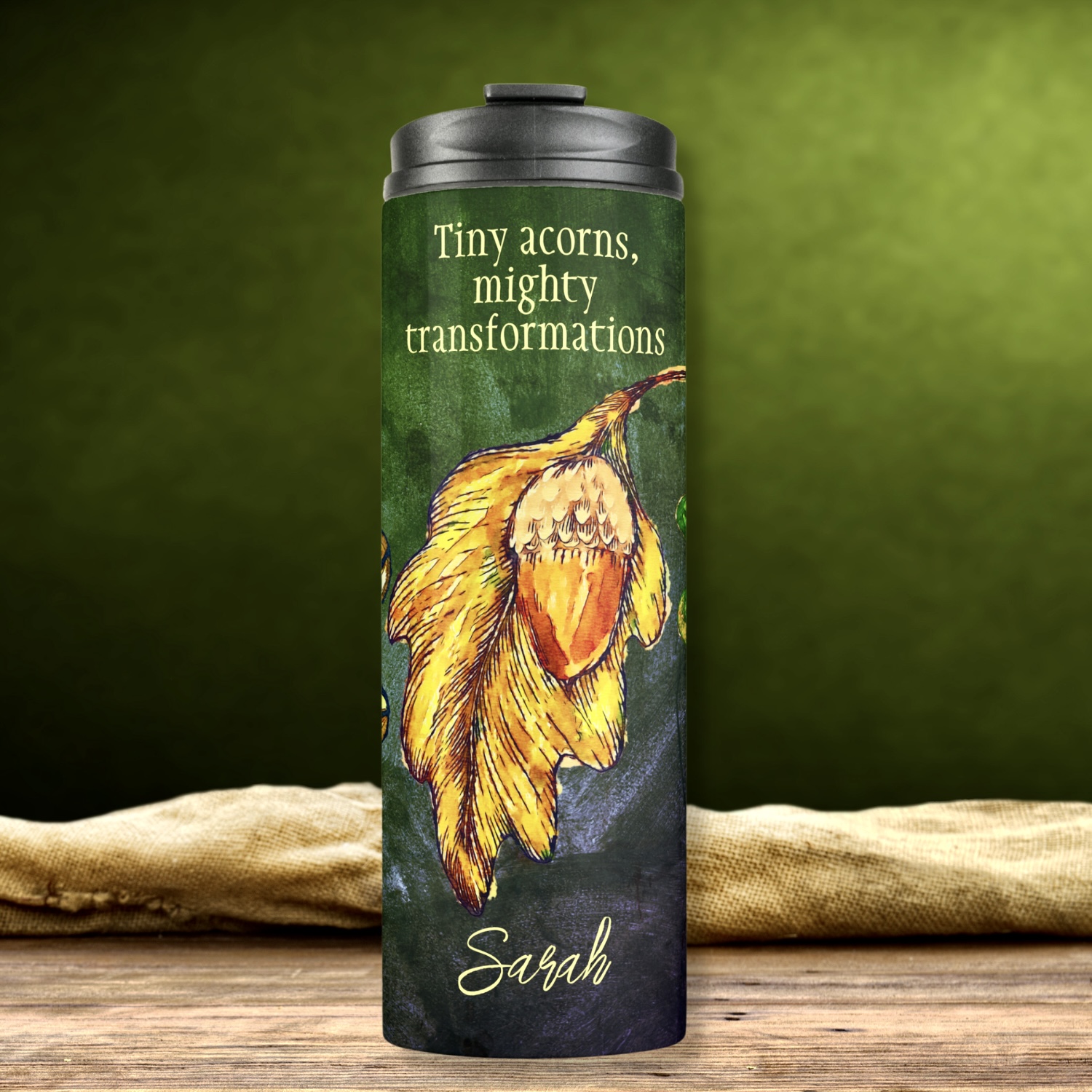 Acorn Fall Colors Inspirational Thermal Tumbler - A bold design featuring lime and golden dry leaves, brightly painted golden and orange acorns, arranged as a collage on a dark hunter green washed-out surface. Customizable quote: 'Tiny acorns, mighty transformations' - personalize with your own quote or name. Two custom fields for personalized messages or names.