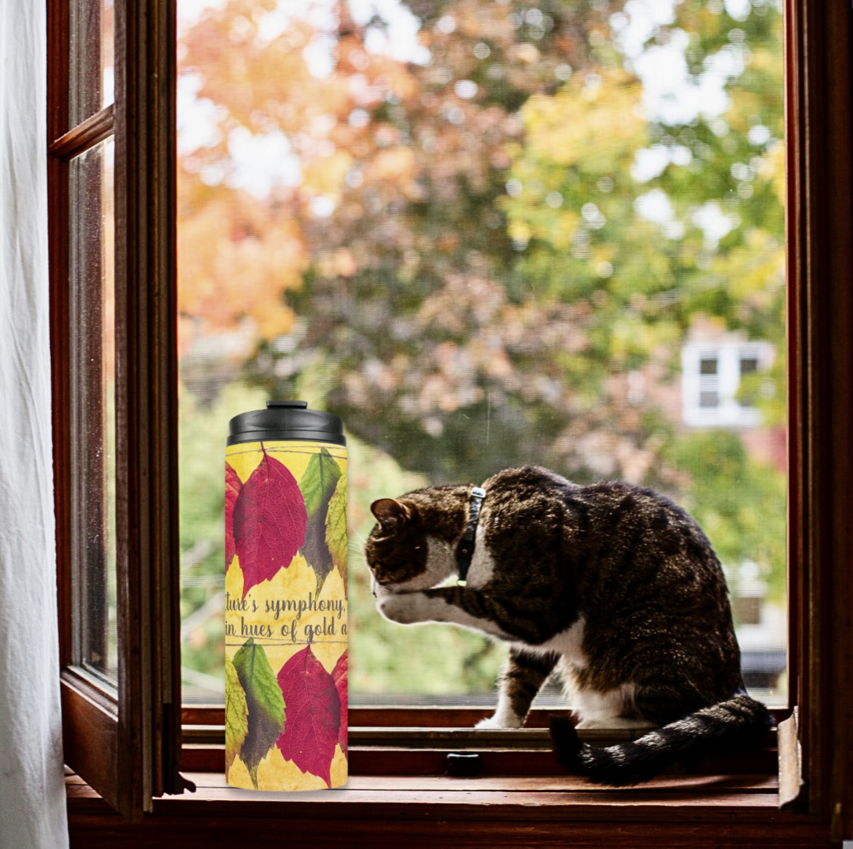 Yellow thermal tumbler with dry red leaves design by an open window, with a cat on the side.