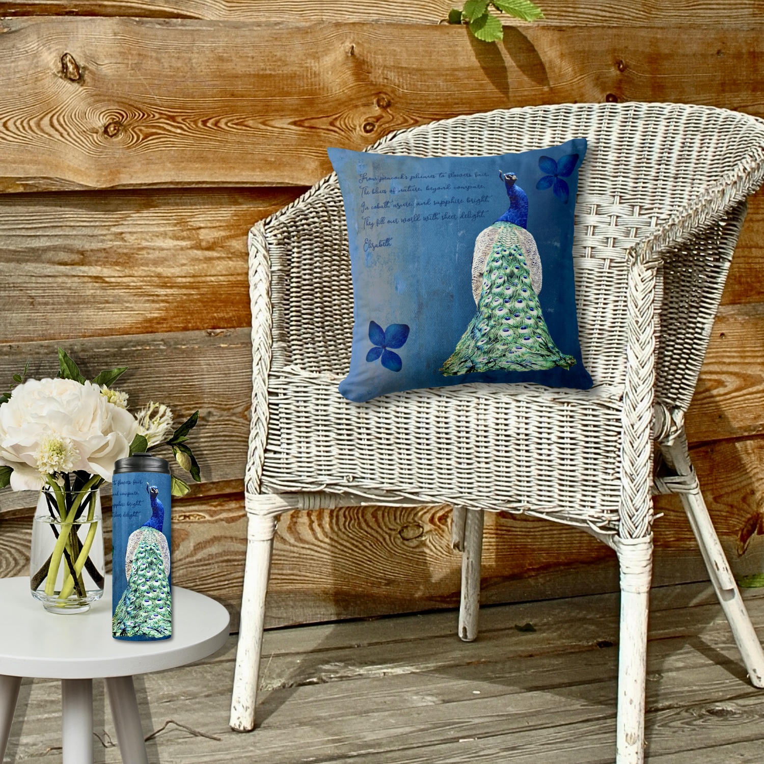 Blue cobalt peacock throw pillow on white rattan chair and tumbler with same design on white table. 
