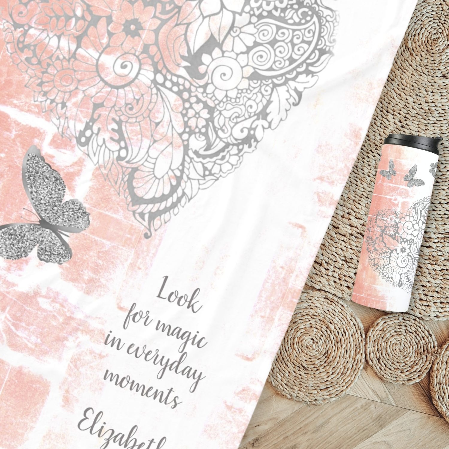 A fleece blanket with a rustic peach design, including a gray heart at its center surrounded by silver butterflies, spread out in a cozy bohemian setting, along with a thermal tumbler with same design.