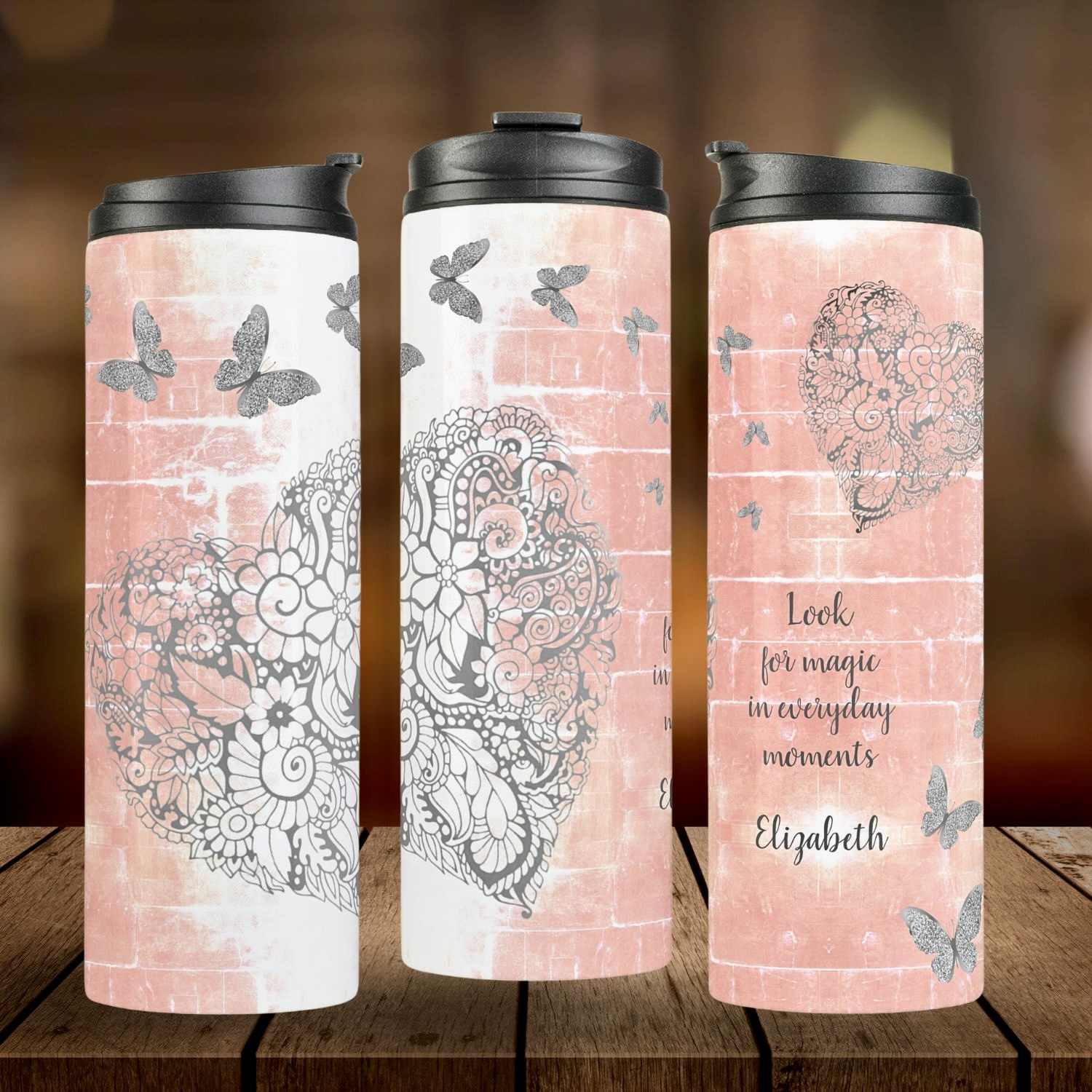 A thermal tumbler with a three-sided design: two sides featuring a rustic peach background with a gray heart and silver butterflies, and the other side displaying an inspiring message.