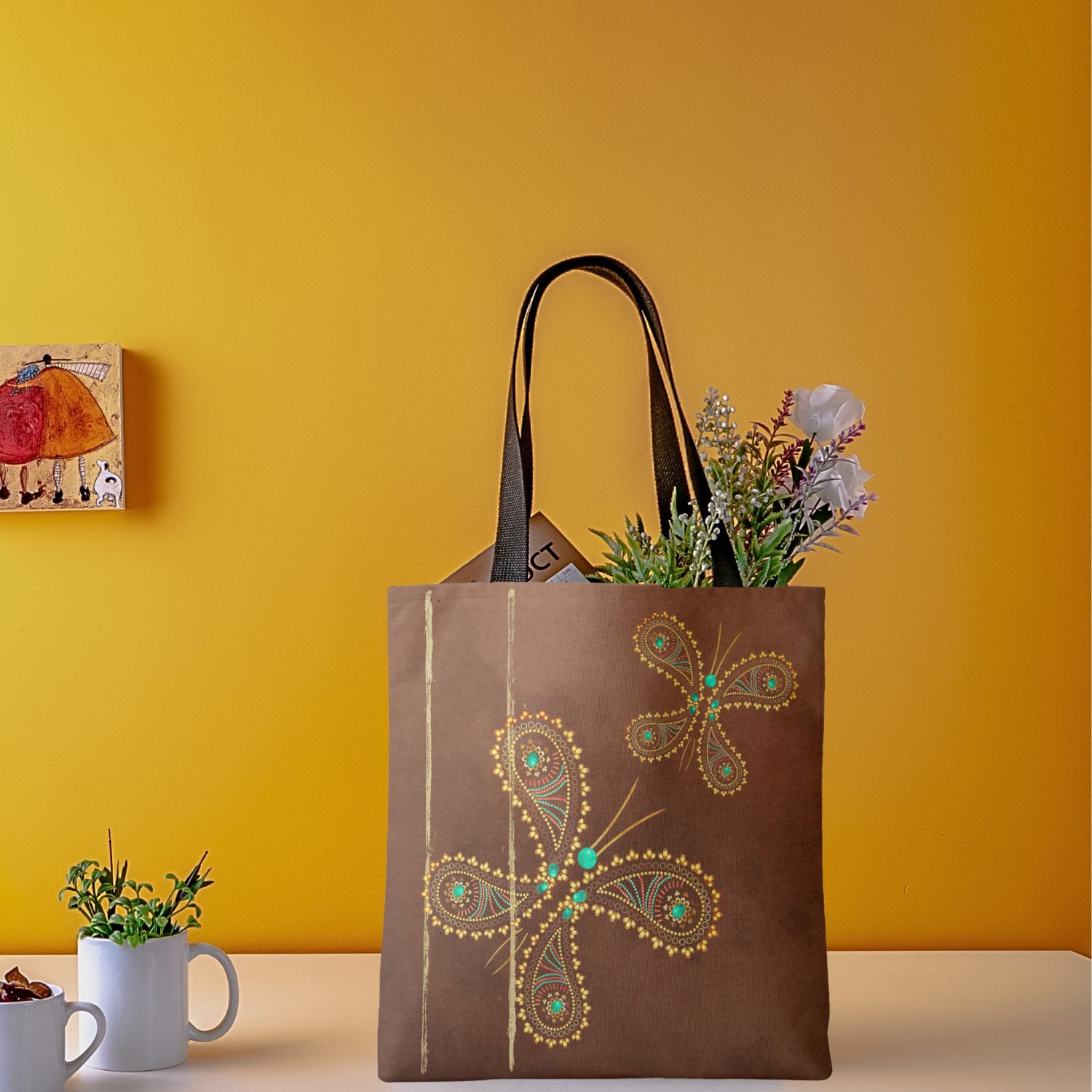 A Brown Turquoise Boho Butterfly Tote Bag is showcased against a contrasting background, with vibrant flowers nestled inside. The tote bag features a charming design of boho butterflies in brown and turquoise hues, adding a touch of whimsy and sophistication to its appearance. The vibrant flowers complement the colorful design, creating a visually appealing contrast. This versatile tote bag is both stylish and functional, perfect for carrying essentials while adding a pop of color to any outfit.