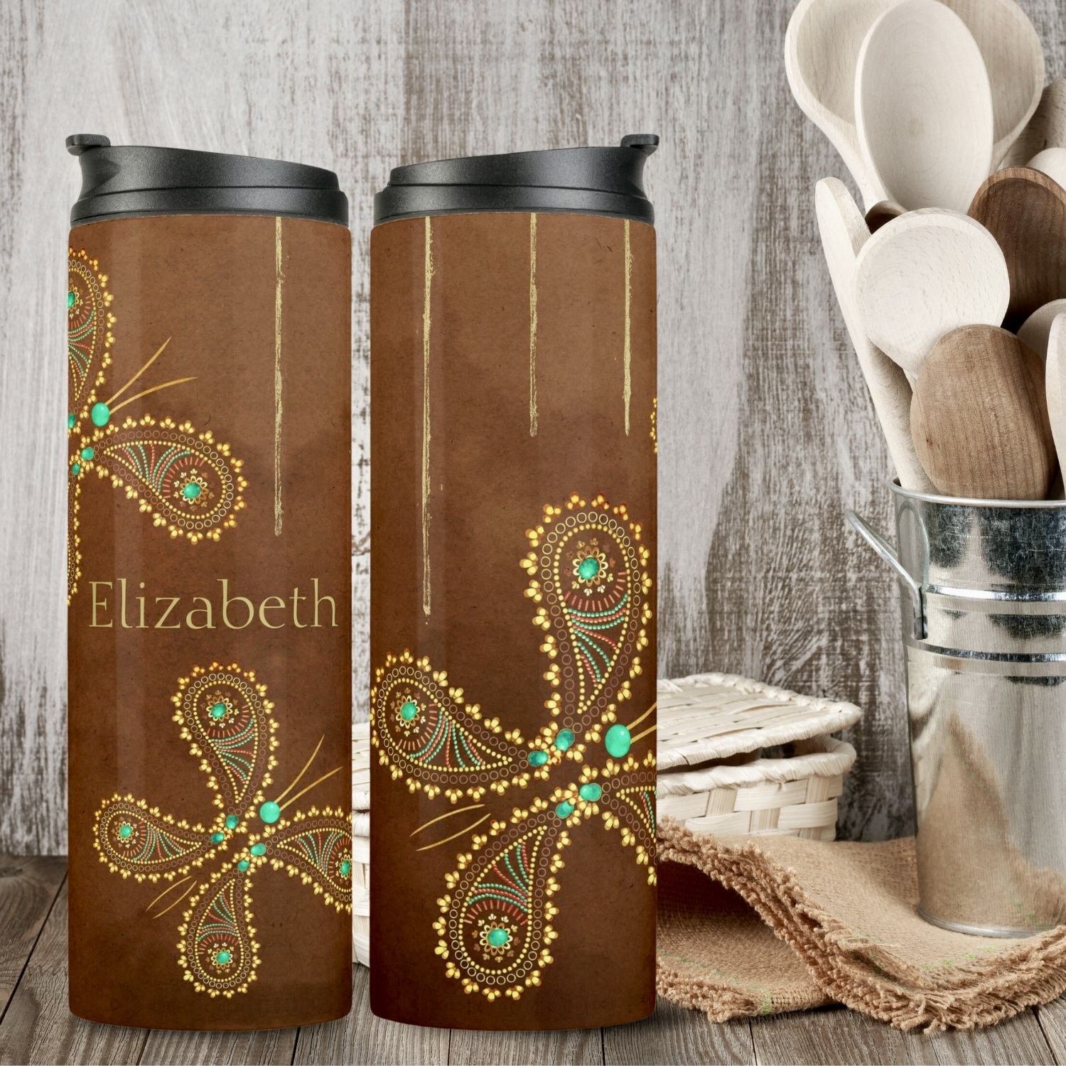 Displayed on a rustic kitchen counter are two similar teal and brown boho butterfly thermal tumblers, showcasing different sides of the tumbler design we created. The tumblers feature intricate butterfly motifs in turquoise and golden hues, adding a touch of whimsy and sophistication. Each tumbler is personalized with a name, creating a unique and thoughtful touch. The image highlights both sides of the tumblers, emphasizing their double-sided design.