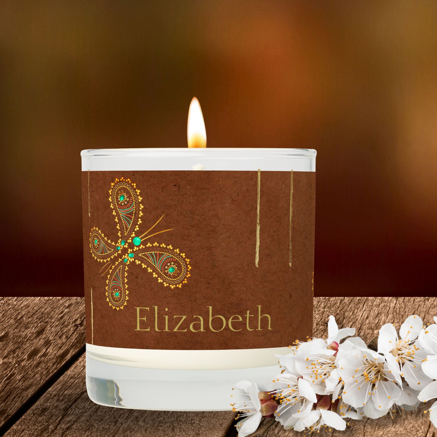 A Teal And Brown Boho Butterflies Scented Candle sits on a rustic surface, accompanied by a white flower. The candle features a charming design of boho butterflies in teal and brown hues, adding a touch of whimsy and sophistication to the ambiance. The rustic surface complements the candle's aesthetic, creating a cozy and inviting atmosphere.