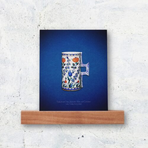 Photo print of a 16th-century Iznik floral tankard, a public domain masterpiece featuring hyacinths, carnations, blue tulips, and red roses. The tankard, influenced by Chinese blue-and-white porcelain, reflects the unique Iznik style from the 1550s and its Ottoman art heritage. The print is displayed in a classic wooden holder, adding a refined touch to the vibrant colors and intricate details of the design.